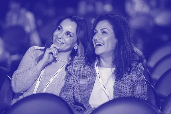 Two women are seated in an audience, smiling and engaged as they watch a presentation. Both women are wearing lanyards around their necks, indicating their participation in an event. The image has a blue tint, enhancing the lively atmosphere of the moment.