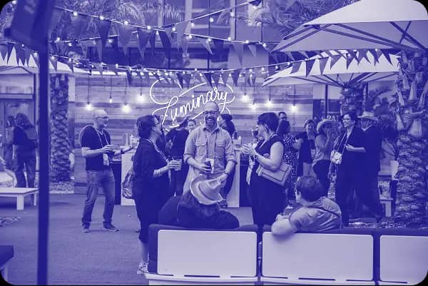 A vibrant outdoor gathering area with a sign that reads "Luminary." Attendees are mingling, some holding drinks, with others seated on white benches. Colorful bunting and string lights add to the festive atmosphere. The scene is lively, showcasing people engaged in conversation under an array of umbrellas, surrounded by palm trees.
