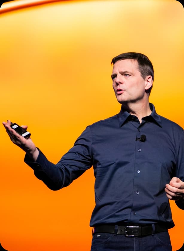A man in a dark shirt gestures with his left hand while holding a remote in his right hand, standing against a bright orange background. He appears to be presenting or speaking in front of an audience.