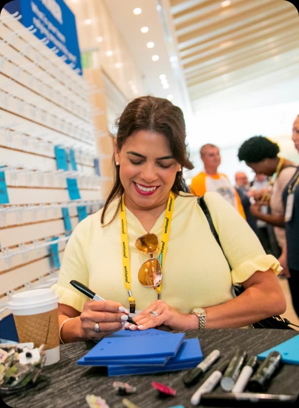 A woman with dark hair and sunglasses writes on a blue notepad at a table with markers and a coffee cup nearby. In the background, people are mingling, and there is a display board with small blue cards.