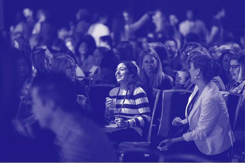 A group of attendees engaged in conversation at a conference. In the foreground, a woman in striped attire smiles while holding a drink. The background is filled with other attendees, some taking notes and interacting with each other. The overall atmosphere is lively and collaborative. The image is presented in a monochromatic color scheme, creating a distinct visual effect.