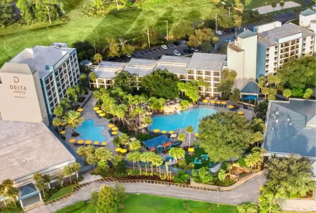 Aerial view of a hotel complex featuring a large pool surrounded by lush greenery and yellow lounge chairs. The Delta Hotel is visible on the left, with additional buildings in the background. The area is well-maintained, showcasing palm trees and landscaped gardens.