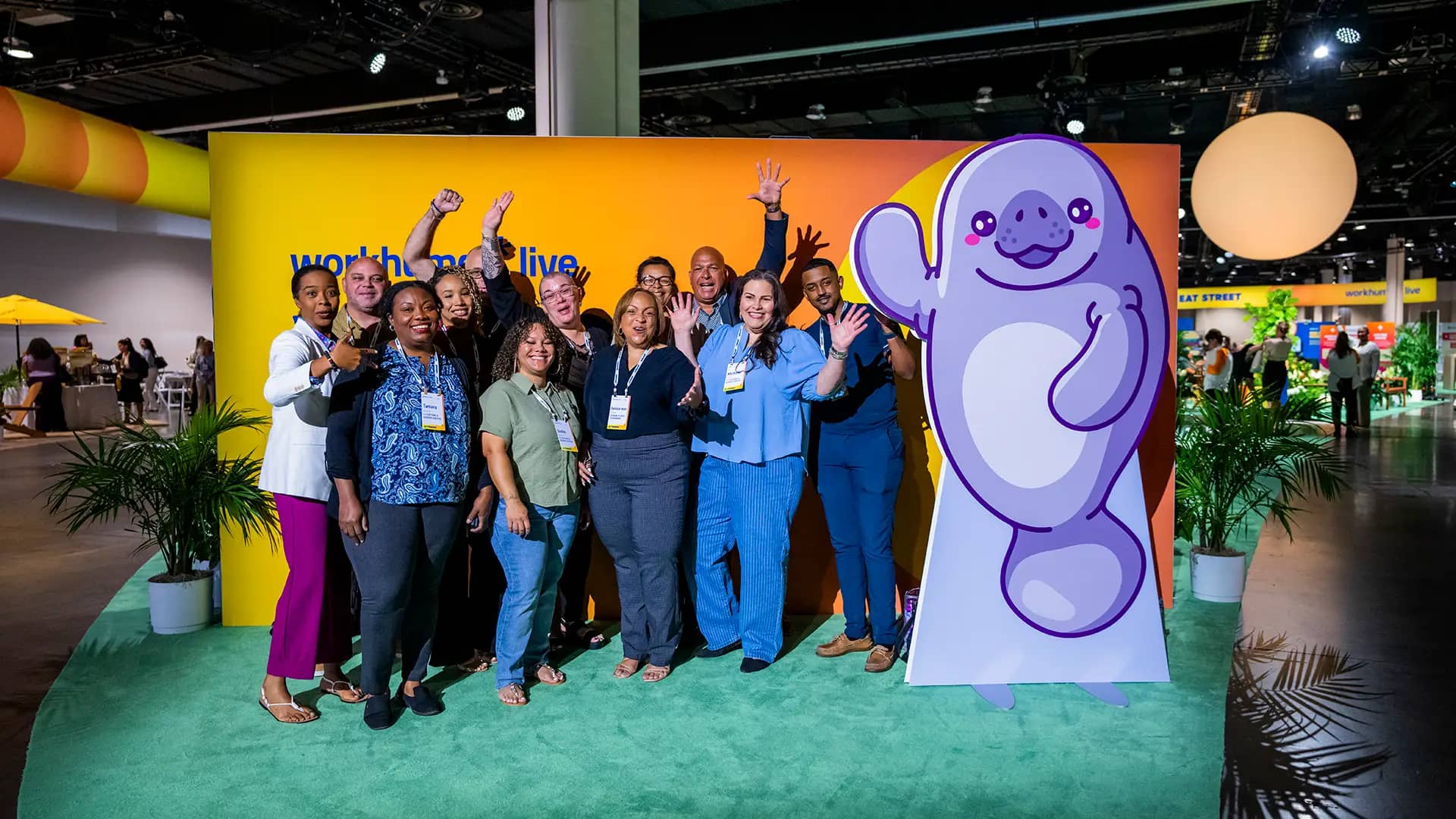 A group of people smiles and poses for a photo in front of a colorful backdrop that features the text "workhuman live." They are gathered around a large, friendly cartoon character resembling a manatee. The setting is vibrant, with plants and event decorations visible in the background. The scene captures a joyful moment at a lively event.