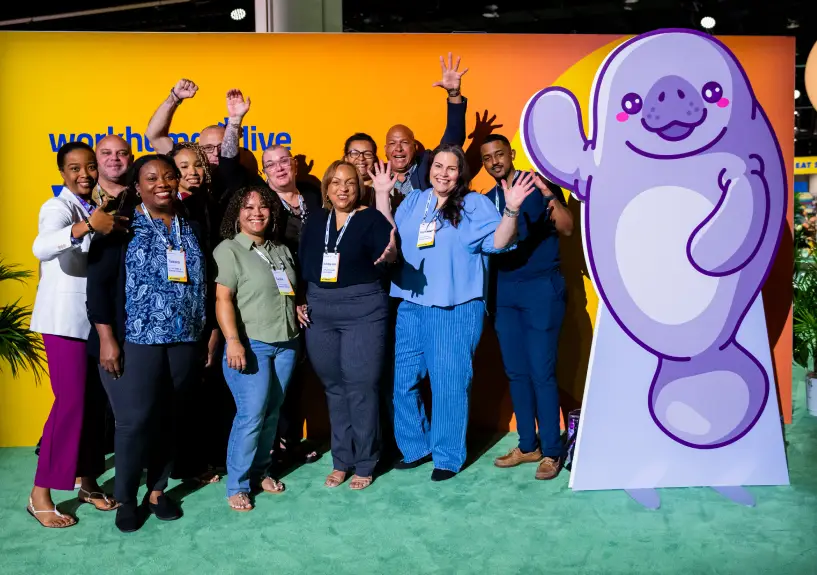 A diverse group of eleven people is posing together in front of a brightly colored background that includes the text "work from home live." They appear to be smiling and waving, and a large cartoon manatee figure is also present beside them. The setting has green carpet and some plants.
