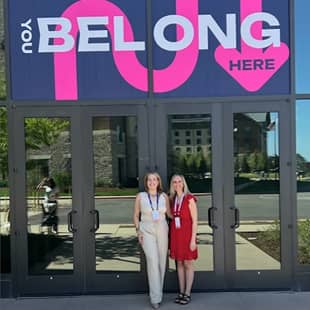 Two women stand together in front of a large glass door featuring a sign that reads "HERE BELONG YOU." The sign uses bright, bold pink letters, and the women are dressed in light and vibrant clothing, smiling at the camera. The background reveals a landscaped area and a building within the vicinity.