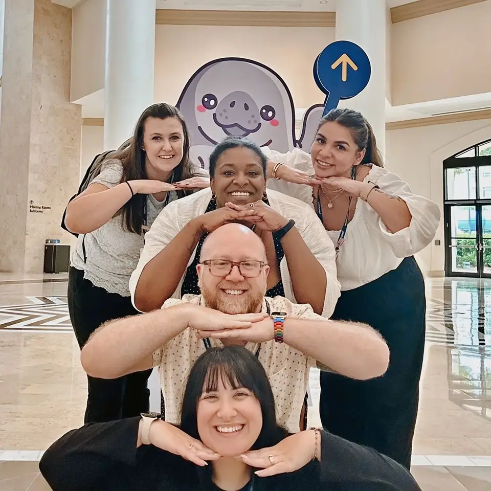 A group of five smiling individuals poses for a photo, with two women standing in the back row and three people in front, playfully placing their hands under their chins. In the background, there is a decorative character resembling a happy manatee, accompanied by a sign with an upward arrow. The setting appears to be a well-lit indoor space, likely part of a conference or event venue.