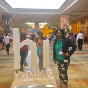 A woman stands beside a large welcome sign that reads "hi HUMANS" in a busy conference setting. The background is filled with attendees engaging and networking, indicating a vibrant atmosphere. Bright lighting emphasizes the openness of the space.