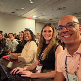 A group of six smiling attendees sit together in a conference setting, facing the camera. They exhibit a sense of camaraderie and engagement, with one person using a laptop. The background shows a conference room with attendees in rows, creating an atmosphere of participation and collaboration during the event.