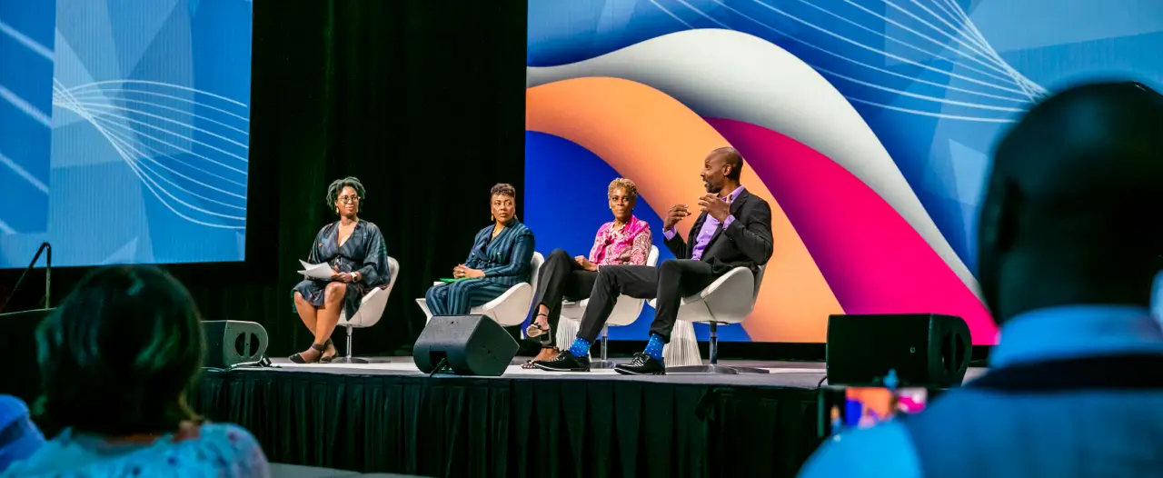 A panel discussion featuring four speakers on stage at a Workhuman event. The panelists are seated in white chairs against a backdrop of colorful curved shapes and lines. The audience is partially visible in the foreground, focusing on the speakers as they engage in conversation. One speaker appears to be making a point while holding a microphone.