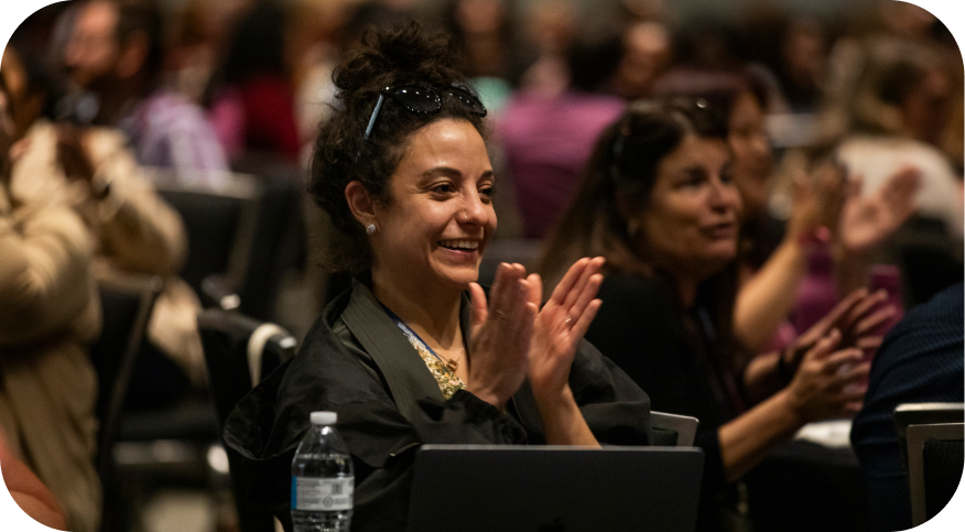 A woman with dark, curly hair and sunglasses on her head is smiling and applauding in an audience at an event. She is seated in front of a laptop, and other attendees can be seen in the blurred background, also engaged in the event.