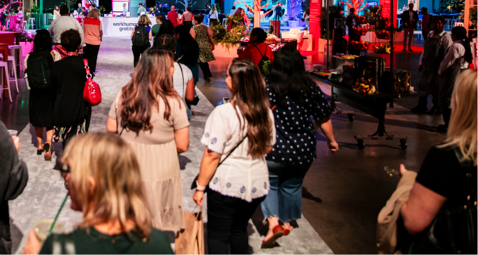 A group of people walk along a bright, colorful exhibition space at a Workhuman event. The attendees, predominantly women, are dressed casually and are engaged in conversation as they move through the venue. Various decorations, including plants and colorful lighting, create an inviting atmosphere, with promotional material for Workhuman visible in the background.