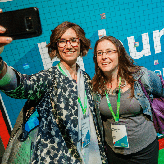 Two women are taking a selfie together in front of a colorful background featuring the word 