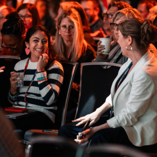 A diverse group of women sit in an audience, engaged and smiling at each other. One woman in the foreground, wearing stripes and large pink earrings, appears to be laughing, while another woman in a white blazer engages with her. Several others in the background hold drink cups, suggesting a lively atmosphere at an event. The attendees are illuminated by warm lighting, enhancing the friendly and inviting mood of the gathering.