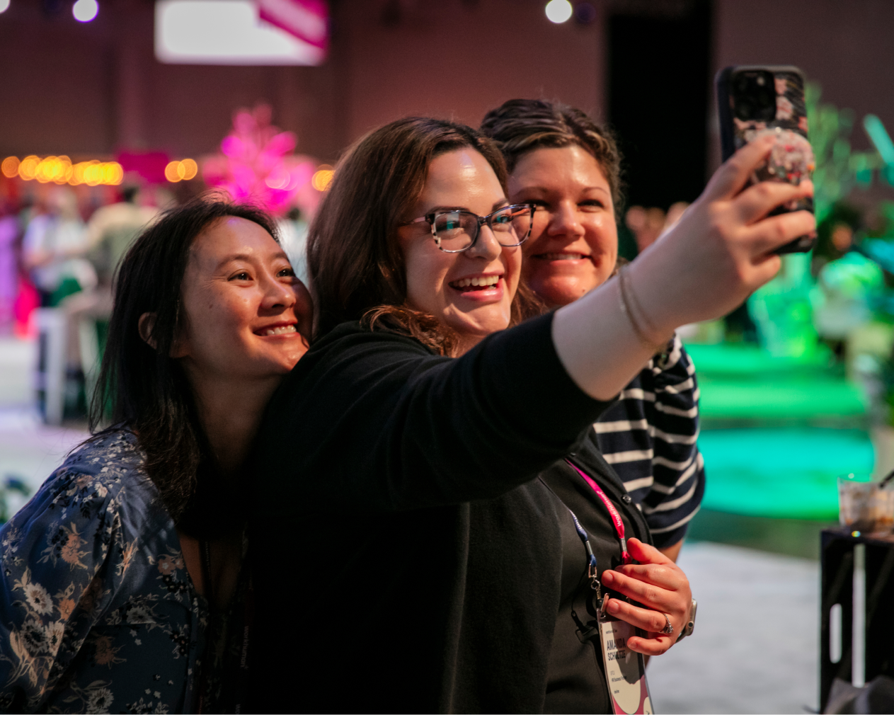 Three women smile while taking a selfie together at a lively event. The background features blurred attendees, decorative lights, and greenery, creating a festive atmosphere. One woman has glasses, another wears a striped shirt, and the third has long hair. They appear joyful and engaged.