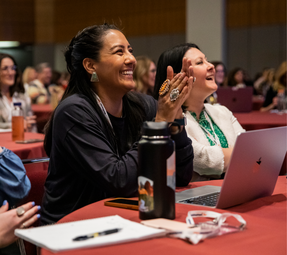 A smiling woman with long, dark hair is clapping and enjoying a presentation in a conference setting. She wears a black top and large, decorative rings. In the background, other attendees can be seen laughing and engaged, with laptops open on the tables and a water bottle in view. The scene captures a lively and positive atmosphere during the event.