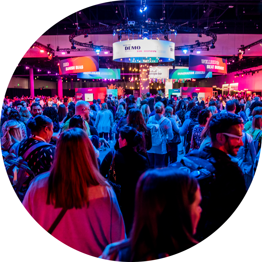 A bustling conference scene at the Workhuman Live event, filled with attendees engaged in conversation. Brightly lit displays feature the words "DEMO," "BANISH BIAS," "INNOVATOR," and "ACCELERATE," showcasing various topics and topics within the space. The atmosphere is vibrant, with a sense of excitement as participants mingle amidst colorful lighting and decorations.