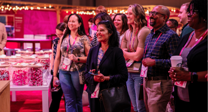 A group of smiling attendees at a Workhuman event stands together in a lively setting, enjoying the experience. They are gathered around a table with colorful treats, dressed in casual attire, and wearing event badges. The atmosphere appears festive, enhanced by warm lighting in the background.