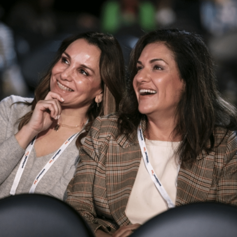 Two smiling women are seated together, engaged in a moment of joy or laughter. They appear to be enjoying an event, with one woman resting her hand on her chin while the other is laughing. Both are wearing lanyards around their necks, suggesting they are attendees at a conference or gathering. The background features blurred outlines of other audience members, enhancing the lively atmosphere.