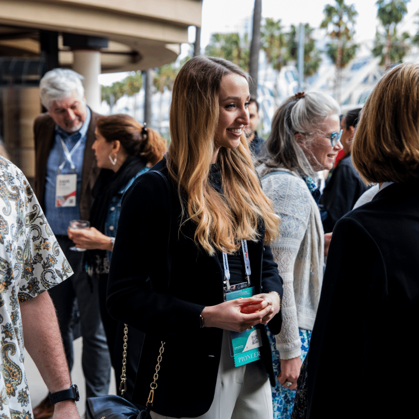 A group of people engaging in conversation outdoors at a conference. A woman with long hair and a black outfit is smiling and holding a red drink, while wearing a name badge that says 