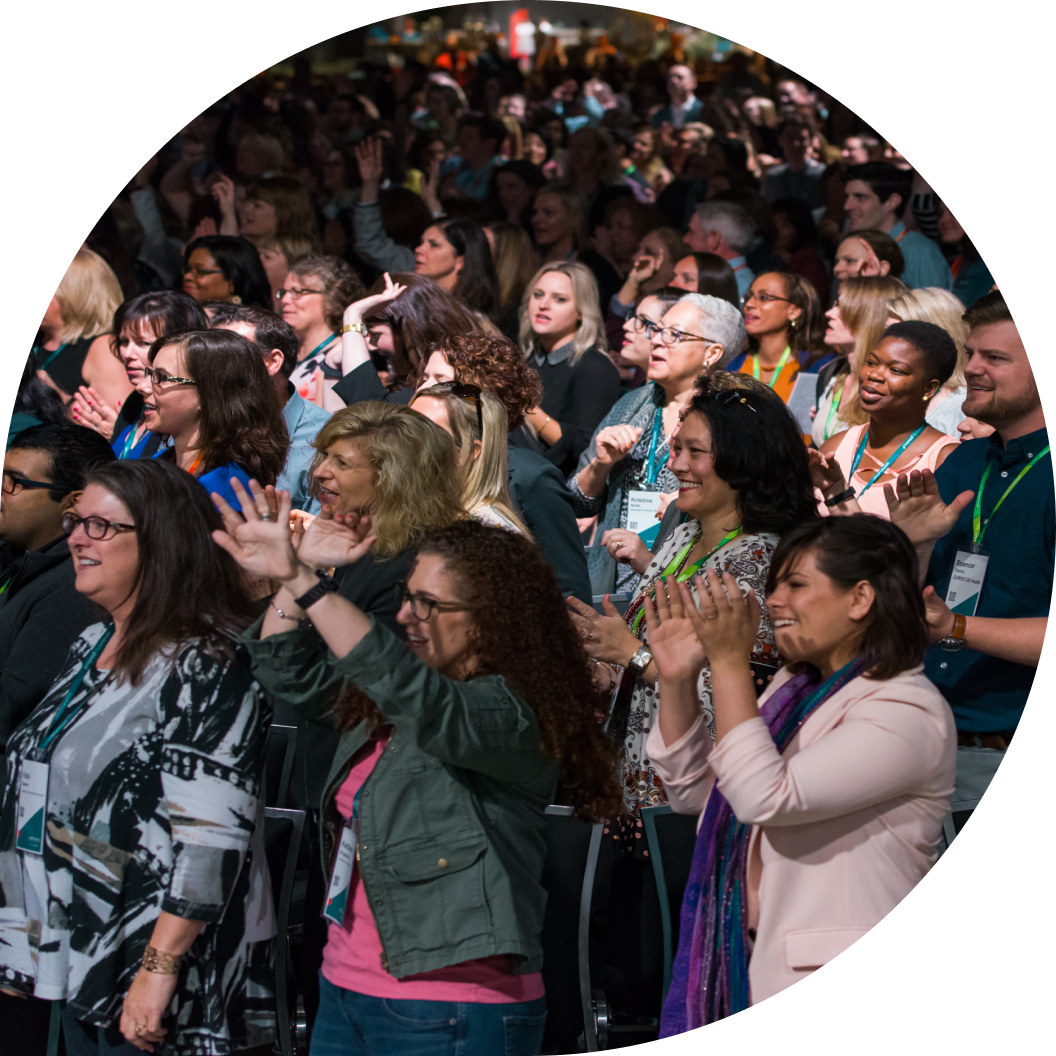 A diverse crowd of attendees at a conference is enthusiastically clapping and raising their hands, smiling as they engage with the event. The atmosphere is lively, suggesting a moment of celebration or excitement. Many participants are wearing badges, and a few are seen in close-up, highlighting their expressions of enjoyment and connection.