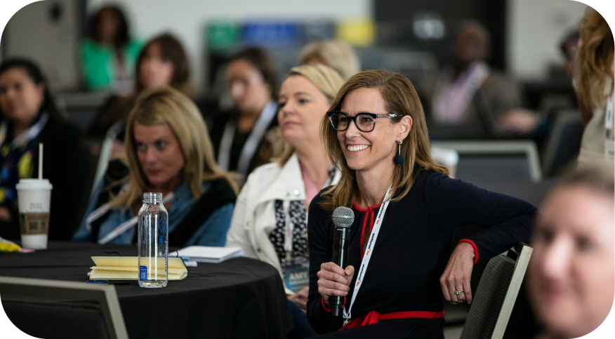 A woman with glasses smiles while sitting at a round table, holding a microphone, and engaging with attendees at a Workhuman event. In the background, several people are seated, showing interest in the discussion. There are various cups and a notebook on the table, creating a lively conference atmosphere.