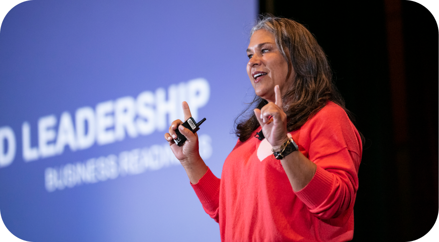 A woman with long hair, wearing a red sweater, gestures while speaking on stage. Behind her is a large screen displaying the text "LEADERSHIP" and "BUSINESS RESEARCH." She appears engaged and energetic, conveying an important message to the audience.