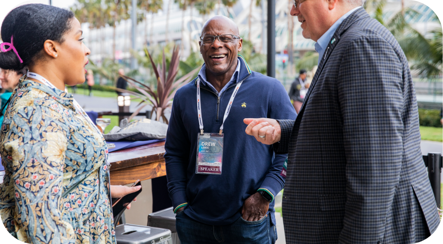 A group of three people engaged in conversation outdoors. One man, wearing glasses and a navy blue sweater, is smiling and appears to be enjoying the conversation while holding a drink. He has a name tag visible that reads "DREW" and "SPEAKER." The two other individuals, a woman with short hair and another man in a checkered blazer, are facing him, contributing to the lively exchange, with plants and outdoor decorations in the background.