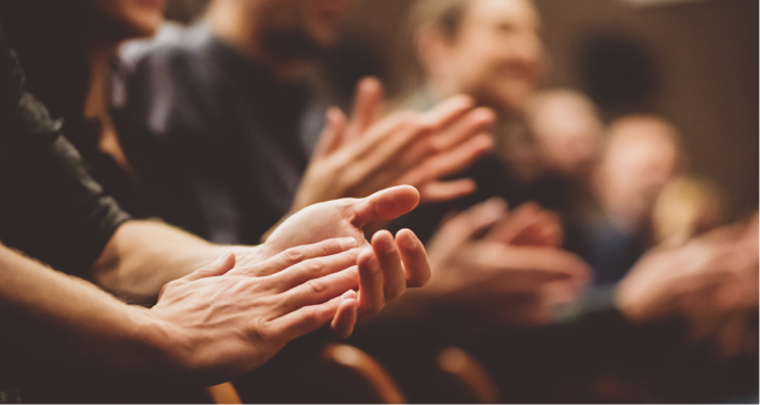 A close-up of several hands clapping in appreciation, with blurred faces of an audience in the background, suggesting a moment of applause or celebration at an event.