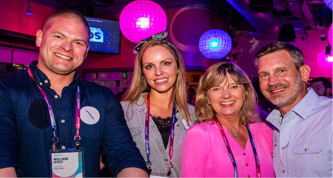 A group of four smiling individuals stand together at a lively event illuminated by colorful lanterns. The first person on the left has short hair and is wearing a dark shirt with a name tag visible. Next to him is a woman with long hair, wearing a black top and a patterned headband. The third individual is a blonde woman dressed in a pink top, and on the right is a man in a light-blue button-up shirt. The atmosphere appears vibrant and cheerful, typical of an engaging social gathering.