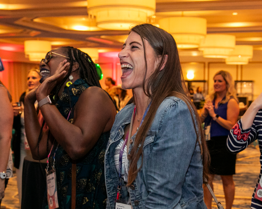 A group of women enthusiastically enjoying an event, with one woman laughing heartily in the foreground. The background features a lively atmosphere, with soft lighting and attendees engaged in conversation, highlighting the joyful ambiance of the gathering.