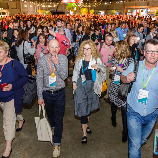 A large crowd at a conference event, with attendees walking through a spacious area adorned with bright lights and decorations. Many individuals are wearing name tags and holding various items, while some are engrossed in conversations. In the background, colorful banners and displays are visible, creating an energetic atmosphere.