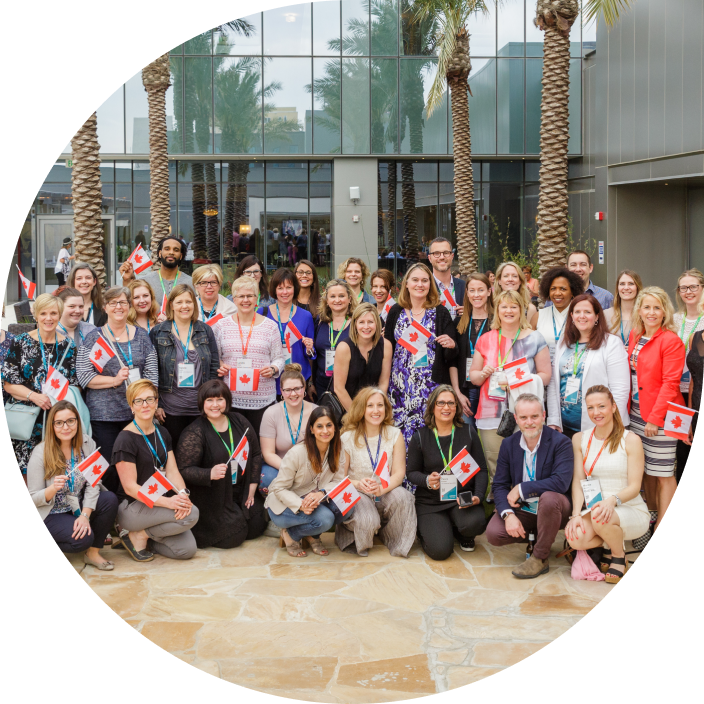 A group photo of diverse individuals gathered outdoors, many holding small Canadian flags. They are smiling and wearing colorful lanyards, standing in front of a modern building with palm trees in the background. The atmosphere appears celebratory and festive.