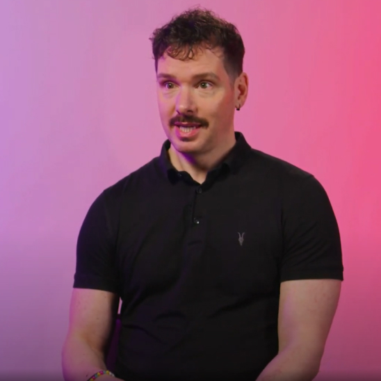 A man with a thick mustache and curly hair sits against a gradient background of pink and purple hues. He is wearing a black polo shirt and appears to be engaged in conversation, looking directly at the camera with an attentive expression.