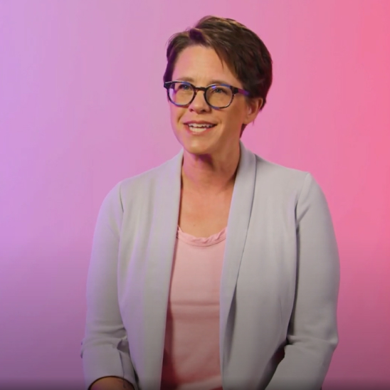 A smiling woman with short brown hair and glasses is seated against a softly lit background that transitions from pink to purple. She is wearing a light purple blazer over a pink top, exuding a warm, approachable demeanor.