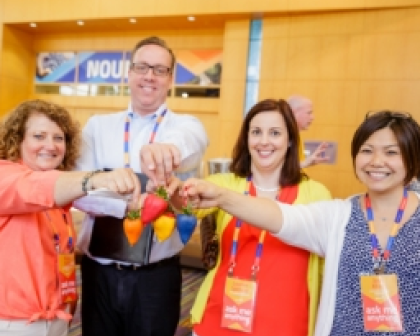 A group of four participants at a conference are smiling and holding colorful hats together in a cheerful display. They are wearing name tags and are positioned in front of a wooden wall with event signage in the background. The atmosphere appears friendly and collaborative, reflecting engagement among attendees.