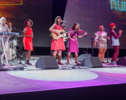 A diverse group of six musicians performing on stage at a Workhuman event. One musician is playing an acoustic guitar, while others are playing keyboard and other instruments. They are dressed in vibrant outfits and are engaging with the audience, creating an energetic atmosphere. The backdrop features colorful lights with the word 