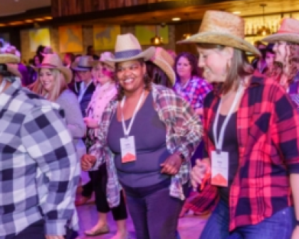 A group of attendees participating in a lively line dance at a Workhuman event, dressed in flannel shirts and cowboy hats. The atmosphere is festive, with colorful lighting enhancing the energetic vibe of the gathering.