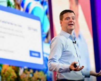 Eric Mosley on stage, engaging the audience while gesturing with his hands. Behind him, there is a presentation slide displayed, featuring text and visual elements related to the topic being discussed.